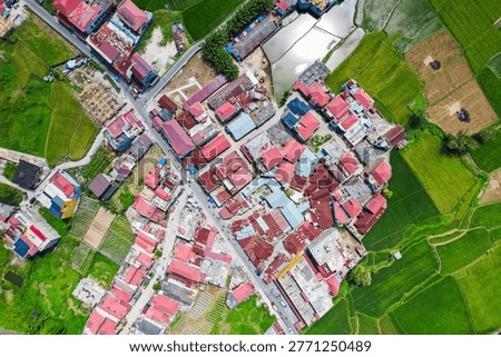 Flat lay drone shot capturing the striking visual contrast between clustered colorful rooftops of a rural settlement and vibrant green farmlands in the peri-urban area of Bukittinggi, West Sumatra, Indonesia.