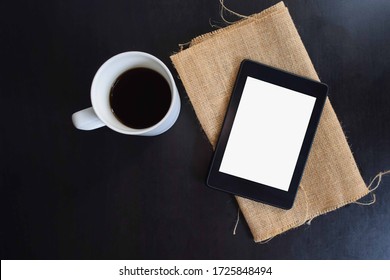 Flat lay of an electronic tablet with a blank screen on a burlap sack and cup of coffee on a black wooden table with copy space. - Shutterstock ID 1725848494