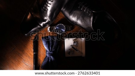 Flat lay of elegant men’s accessories including polished black leather dress shoes, a wristwatch, cufflinks, a bow tie, and a pen arranged on a wooden surface. Dramatic lighting creates a sophisticated and luxurious atmosphere, ideal for concepts related to fashion, business style, formal events, and men’s grooming.     