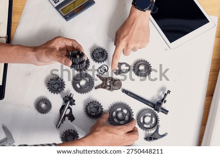 Flat lay of engineering workspace. Two professionals sorting mechanical automotive components and checking blueprint data on tablet. Mechanical engineers team discussing industrial mechanism.