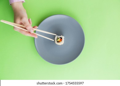 Flat Lay Of Female Hand Holding Chopsticks And One Sushi Roll On Plate Isolated On Green Background.