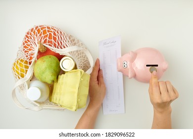 Flat Lay With A Female Hand Putting A Coin In A Piggy Bank, String Bag Full Of Groceries - Eggs, Pear, Milk, Pumpking, Lemon, Yogurt And Receipt On White Background.