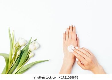 Flat Lay Female Hands Apply Cosmetic Skin Care Cream Next To Bouquet Of Fresh Delicate Tulip Flowers On White Background.