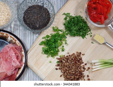 Flat Lay Of Fresh Ingredients Of A Wild And Brown Rice Dressing Recipe Includes Prosciutto, Pecans, Green Onions, Parsley And Pimento. The Ingredients Are Scattered On A Wood Cutting Board And Table.