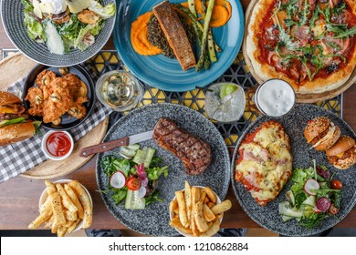  Flat Lay Of Fried Chicken, Caesar Salad, Grilled Steak, Chips, Parmigiana, Pizza, Burgers, Sliders, Grilled Salmon, Beer And Wine On A Wooden Table.