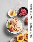 Flat lay of greek natural yogurt with oat granola or muesli, peach, raspberry and honey in a bowl on a light background with fresh fruits and berries. Healthy and nutritious breakfast concept.