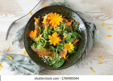 Flat Lay Of Green Plate With Arugula, Orange, Sesame Seeds And Edible Marigold Flowers Salad On Light Table, Vegetarian Food Concept