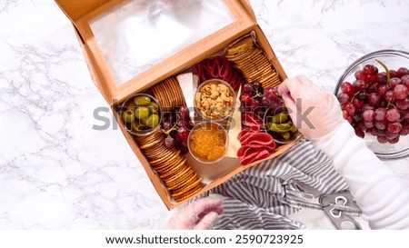 Flat lay. Hands are meticulously adding fresh red grapes to a bowl, complementing a beautifully arranged charcuterie box brimming with a variety of cheeses, olives, and cured meats, set against a