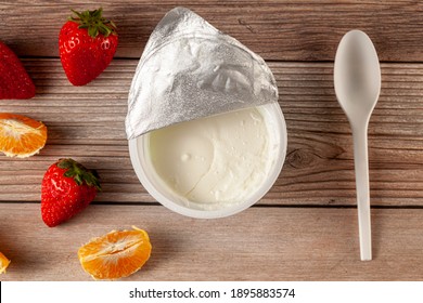 Flat Lay Image Of A Store Bought Plastic Cup Of Plain Low Fat Greek Yogurt With The Aluminum Foil Seal Peeled Off. It Is On Wooden Background With A Plastic Spoon And Fresh Fruits. A Diet Idea Concept