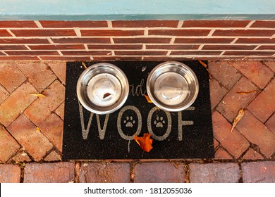 Flat lay image of two metal plates filled with drinking water for stray animals that are placed on a black doormat with "WOOF" written on it and dog paws figures in the middle. Animal friendly concept - Shutterstock ID 1812055036