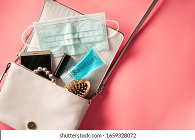 Flat Lay Of Ivory Leather Woman Bag Open Out With Face Mask ,sanitizer Hand Gel To Protect From Coronavirus Or COVID-19, Lipstick, Accessories And Smartphone On Pink Background With Copy Space