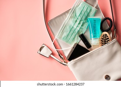 Flat Lay Of Ivory Leather Woman Bag Open Out With Face Mask ,sanitizer Hand Gel To Protect From Coronavirus Or COVID-19, Lipstick, Accessories And Smartphone On Pink Background With Copy Space