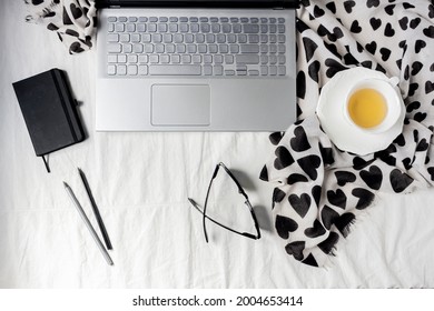 Flat lay of laptop, notebook, black sun glasses, scarf, black and grey pencils and a cup of chamomile tea on a white cotton fabric. Top view. Work at home concept.