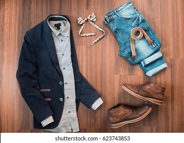 Flat Lay Of Mens Casual Clothes Set Such As Blue Suit With Patches, Jeans, Checkered Bowtie, Belt And Shoes On Brown Wooden Background. Top View.