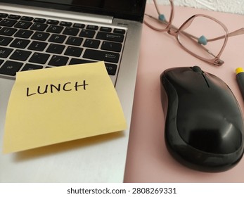 Flat lay of a modern pink workspace showing human traces during lunch break. A yellow sticky note with handwritten LUNCH text placed on a silver laptop, with eyeglasses, wireless mouse and a pen on the side. Concept of taking a lunch break, away from desk, work life balance and empty workplace.