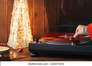 Flat lay Noel greeting card young woman in a red knitted winter pullover puts a red vinyl record on a turntable, a stylus a needle and listens to music a glowing Christmas tree and a cozy New Year's - Powered by Shutterstock