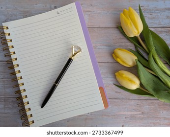 Flat Lay Of A Notebook And Pen On A Wooden Surface With Three Yellow Tulips And Green Leaves With White Space