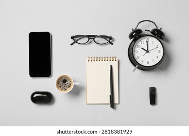 Flat lay of office essentials including a smartphone, coffee, notebook, alarm clock, and glasses on a light gray background
