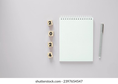 Flat lay of an open blank white page of a notepad with a pen on a side and numbers 2024 on wooden cubes, light lilac background with copy space