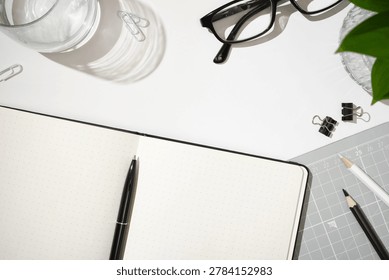 Flat lay of open dotted notebook, black pen, glasses, paper clips, binder clips, pencils, and cutting mat on white desk background. Copyspace