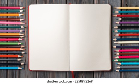 Flat lay of open empty blank notebook between colored pencils in row on grey wooden table. Back to school, art, drawing, stationary, education, office supplies. Top view, copy space, place for text.