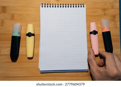Flat lay of open grid notebook with colorful markers. Hand holding pink highlighter on wooden background. Top down view of office supplies for studying, planning or creative work. Blank copy space.