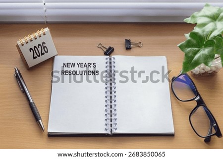 Flat lay of open notebook title NEW YEAR'S RESOLUTIONS on wooden desk with copy space, 2026 calendar, pen, glasses, binder clips, and green plant; goal setting, New Year planning, productivity concept