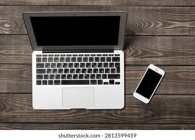 Flat lay of an open silver laptop and a modern smartphone with a blank screen placed on a dark brown wooden textured surface for professional workspace concept