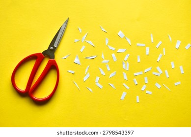 Flat lay of a pair of red-handled stainless steel scissors alongside numerous tiny scattered white paper scraps on a vibrant yellow paper surface.