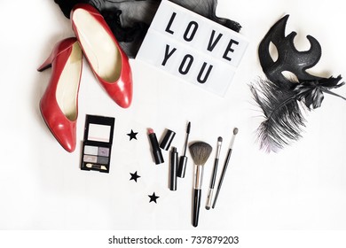 Flat lay of party preparation accessories. top veiw: red high heels shoes, cosmetics and brushes, black mask and I love you note on a word table