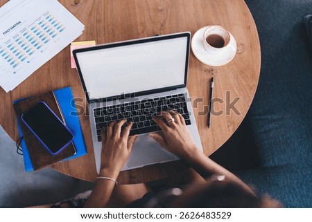 Flat lay perspective of hands on keyboard working remotely in café, with smartphone, sticky notes, coffee cup and project charts on wooden table indicating busy freelancer lifestyle
