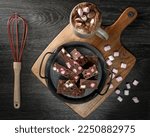 A flat lay photo of rocky road chocolate fudge, on a vintage serving tray and board. A cup of hot chocolate and pink and white marshmallows and a red whisk are next the fudge.