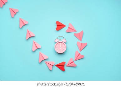 Flat lay with pink and red paper airplanes around a pink clock on a blue background. Time flies concept with paper planes flying around the alarm clock