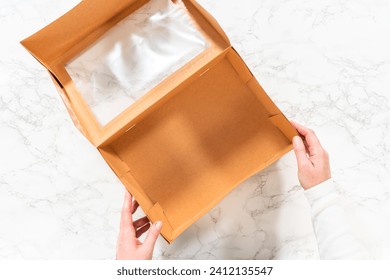 Flat lay. In the process of assembling, a woman expertly curates this charcuterie box, showcasing sliced meat, cheese, crackers, and grapes, all neatly arranged and packaged in a brown gifting box.
