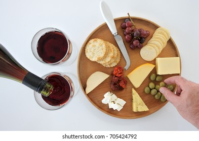 Flat Lay Of Red Wine Poured Into Two Wineglasses And Mans Hand Selecting An Olive  From A Cheese Platter. Food Background. No People. Copy Space