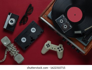 Flat lay retro media and entertainment. Vinyl record player with vinyl record, film camera, video cassette, audio cassette, gamepad, handset on red background. 80s. Top view