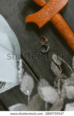 A flat lay shot of a silver and a gold wedding ring placed on a dark wooden surface next to a traditional Malay wooden kris (Keris) handle.