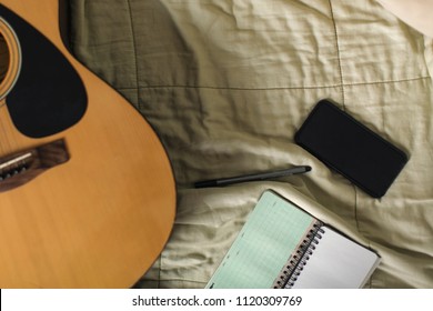 Flat lay of smartphone, notepad, pen, and guitar on  bed