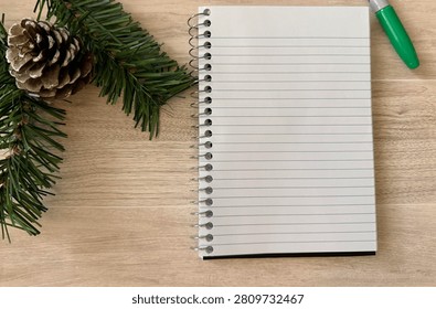 Flat Lay Of Spiral Bound Lined Notebook Green Capped Marker And Artificial Snow Capped Pine Cone And Evergreen Needles As Christmas Decor On Light Wooden Desk 