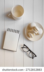 Flat lay of a spiral notepad, reading glasses, empty coffee mug and plate of white macarons on a clean white wooden desk, suggesting a quiet work break or planning session