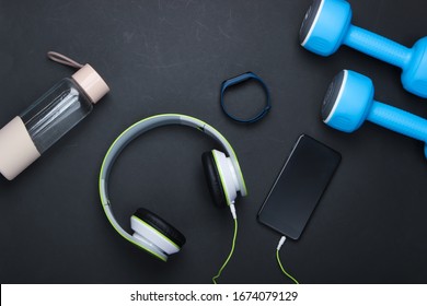 Flat Lay Sports And Fitness Composition. Modern Gadgets. Dumbbells, Headphones With Smartphone, Smart Bracelet, Water Bottle On Black Background. Top View