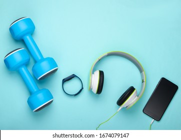 Flat Lay Sports And Fitness Composition. Modern Gadgets. Dumbbells, Headphones With Smartphone, Smart Bracelet On Blue Background. Top View