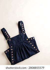 Flat lay studio shot of dark blue denim dungaree dress with copper snap buttons on off white background
