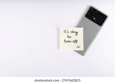 A flat lay studio shot featuring a grey smartphone with a yellow sticky note on its screen. The note reads, 