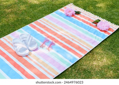 Flat lay of summer essentials including white sandals, pink sunglasses, and pink flowers on a colorful striped beach towel laid out on green grass.