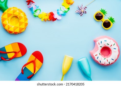Flat lay of summer vibes concept with colorful pool party items, funny sunglasses, cocktail glasses, pineapple and donut inflatable drink holders, flip flops and flower necklaces on blue background