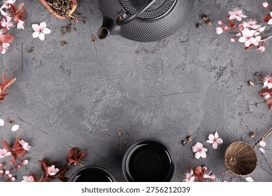 Flat lay tea composition with cast iron teapot, cups, tea strainer, loose tea leaves, and cherry blossoms forming a decorative frame on a dark stone background with large copy space.