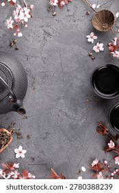 Flat lay tea composition with cast iron teapot, cups, tea strainer, loose tea leaves, and cherry blossoms forming a decorative frame on a dark stone background with large copy space.