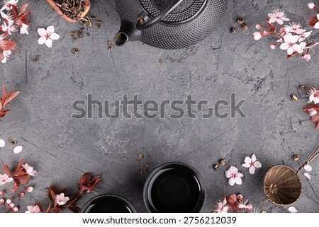 Flat lay tea composition with cast iron teapot, cups, tea strainer, loose tea leaves, and cherry blossoms forming a decorative frame on a dark stone background with large copy space.