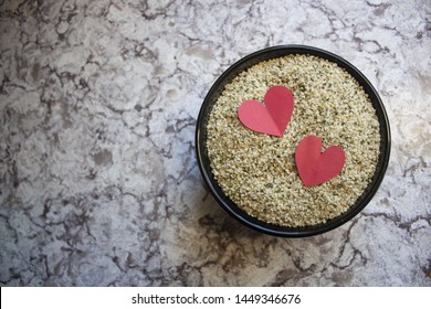 Flat Lay Top Down Shot Of A Bowl Of Hemp Hearts With Two Red Paper Hearts Laid On Top On A Marble Counter Top